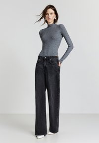 Frau mit langen braunen Haaren, die einen eng anliegenden grauen Rollkragenpullover, weit geschnittene schwarze Jeans, weiße spitz zulaufende Schuhe und silberne Creolen trägt.