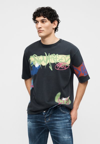 Νέος άνδρας με σγουρά μαλλιά φορώντας μαύρο T-shirt με γραφικά σε στυλ γκράφιτι και φθαρμένα μπλε τζιν, στέκεται με τα χέρια στις τσέπες.