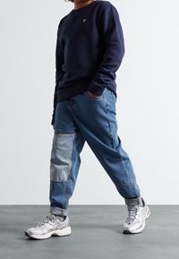 Granatowy sweter z małym żółtym logo, w połączeniu z dżinsami w stylu patchworkowym w jasnoniebieskim i ciemnoniebieskim kolorze oraz szarymi butami sportowymi z teksturowaną podeszwą.