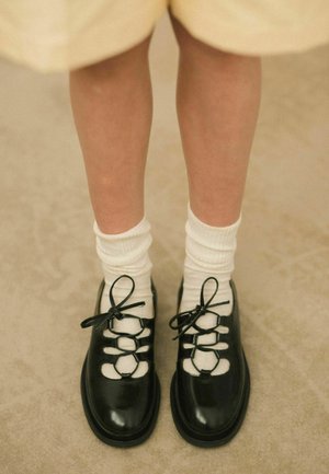 Chaussures noires en cuir verni avec des accents découpés blancs et un design à lacets, accompagnées de chaussettes blanches côtelées, présentées sur un fond neutre.