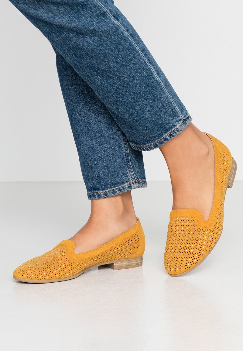Jambes en jean bleu portant des chaussures plates jaunes perforées à enfiler, debout sur un sol blanc lisse.