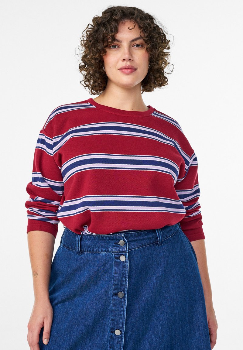 Červený sveter s bielymi a modrými horizontálnymi pruhmi, okrúhlym výstrihom a dlhými rukávmi, kombinovaný s rifľovou sukňou s prednými gombíkmi.