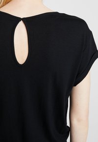 Rückansicht einer Person in einem schwarzen Kurzarmhemd mit einem Schlüsselloch-Ausschnitt und Knopfverschluß im oberen Rückenbereich.