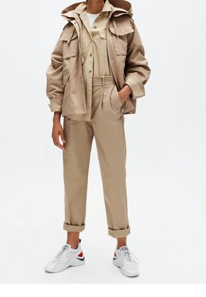 Persona con chaqueta de trabajo beige, pantalones de talle alto a juego con puños, zapatillas blancas con detalles rojos y negros, de pie con una mano en el bolsillo.