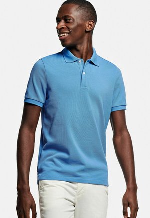 Lachende man die een lichtblauw poloshirt met korte mouwen en witte broek draagt, kijkend naar links tegen een effen witte achtergrond.