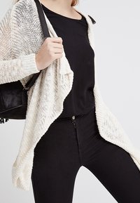 Cardigan en tricot ouvert de couleur crème, présentant une coupe ample, un motif texturé et des manches tombantes. Associé à un top noir et un jean.