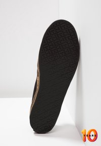 Schwarze Sneaker-Sohle mit texturiertem Profilmuster. Die Seite hat ein metallisches goldfarbenes Akzent. Oben ist überwiegend schwarz, minimalistisches Design.