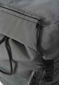 Sac à dos gris avec un tissu texturé à rayures verticales, fermoir à cordon et accents matériels noirs. Bandoulières latérales réglables incluses.