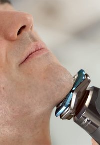 Rasoi elettrico Philips di colore grigio metallizzato e blu, con una testa arrotondata vicina al mento di un uomo, che mette in mostra il suo design elegante e la superficie texturizzata.