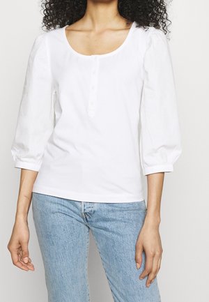 Femme portant un haut blanc boutonné avec des manches trois-quarts bouffantes et un jean bleu clair, debout devant un fond uni.