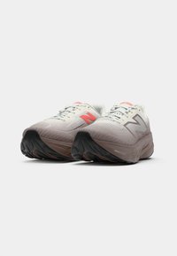 Une paire de chaussures de course New Balance beiges et brun clair avec de grosses semelles noires et des logos orange et gris sur un fond blanc.