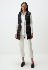 Schwarze Steppweste mit tailliertem Design, Reißverschluss vorne und schmaler Taille; getragen über einem weißen Hemd und kombiniert mit weißen Cropped-Hosen.