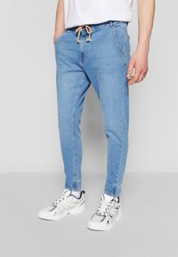 Svetlo modri jeans hlače jogger stila z stranskimi žepi, elastičnim pasom z vrvicami in ožjimi manšetami na gležnjih. Obute imajo bele superge.