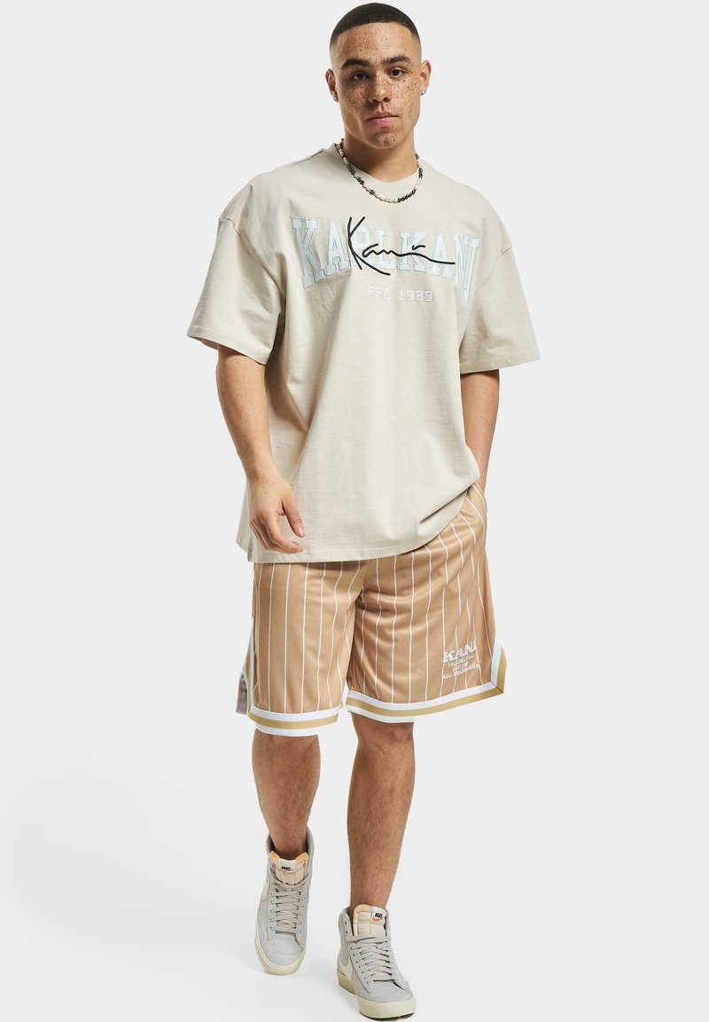 T-shirt en coton beige avec imprimé graphique, associé à un short beige rayé avec des accents blancs. Le modèle porte des baskets montantes grises.