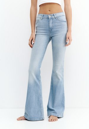 Kvinde iført lyseblå high-waist bootcut-jeans og en hvid cropped top, stående barfodet mod en enkel hvid baggrund.