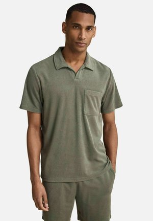 Mann trägt olivgrünes kurzärmeliges Hemd mit Kragen und Brusttasche sowie passende Shorts, steht mit einer Hand in der Tasche.