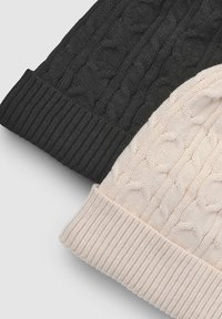 Dwie dziergane czapki z daszkiem, jedna czarna z wzorem warkoczowym, druga kremowa o podobnym wzorze. Obie mają prążkowane obrzeża.