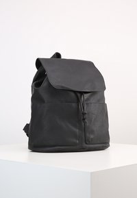 Sac à dos en cuir noir avec rabat avant, fermeture à cordon, et deux poches avant, posé sur un piédestal blanc contre un fond uni.