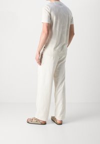 Ljusbeige kortärmad t-shirt med ett subtilt mönster, kombinerad med löst sittande vita byxor och ljusbruna sandaler mot en neutral bakgrund.