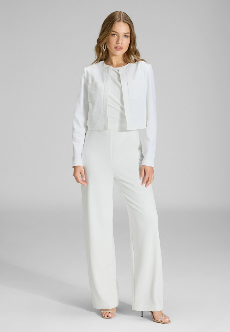 Veste blanche courte avec deux poches, associée à une combinaison blanche avec pantalon large. Texture lisse, design minimaliste, accents argentés sur les chaussures.