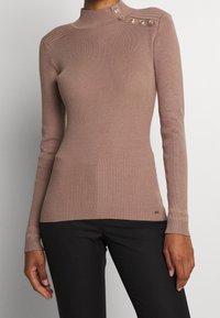 Harmaat ribattiset turtleneck-paita pehmeässä vaaleanpunaisessa värissä, joka istuu hyvin vartalon myötäisesti. Paidassa on koristeelliset olkatoppaukset ja pitkät hihat, jotka on yhdistetty mustiin housuihin.
