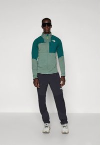 Fleecevest met groene en teal gekleurde delen, voorzien van een rits, gecombineerd met zwarte broek. Accessoires zijn onder andere een zonnebril en beige wandelschoenen.