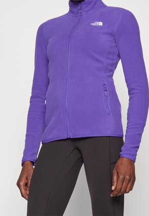 Vrouw draagt een nauwsluitend paarse fleecejas met ritssluiting en het logo van The North Face, gecombineerd met zwarte leggings.