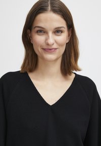 Haut en tricot gaufré noir avec un design à col en V, doté de manches légèrement tombantes et d'une finition texturée. La teinte de peau est neutre.