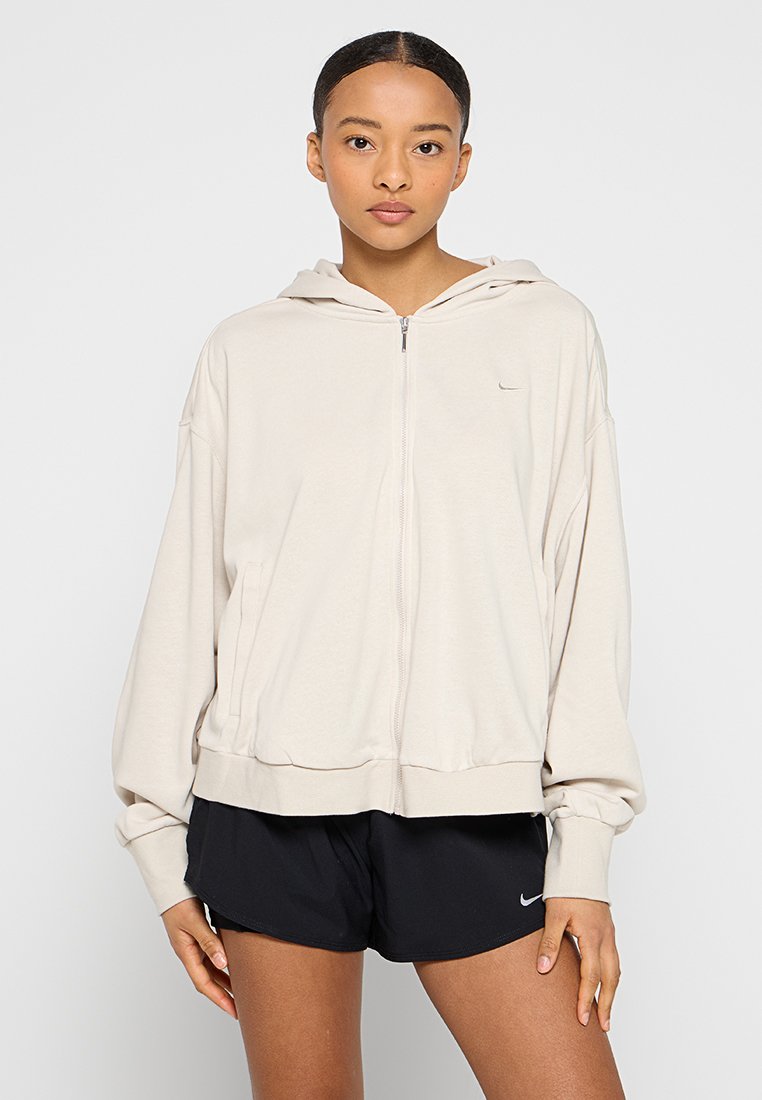 Felpa con cappuccio di un beige chiaro, con zip frontale, vestibilità oversize, con polsini e orlo a coste, dotata di un piccolo logo sul lato sinistro del petto. Abbinata a pantaloni corti neri.