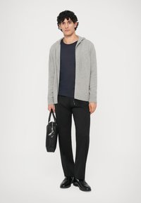 Γκρίζα πλεκτή κουκούλα πάνω από μια ναυτική μπλούζα, σε συνδυασμό με μαύρο παντελόνι και μαύρα παπούτσια. Κρατώντας μια μαύρη τσάντα με φερμουάρ και χερούλι.