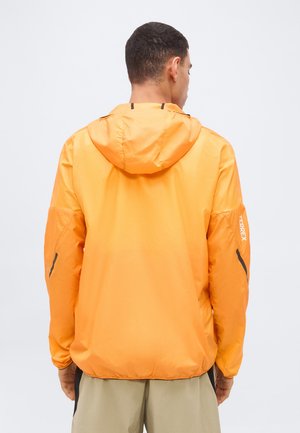 Άνδρας με πλάτη γυρισμένη, φοράει φωτεινό πορτοκαλί μπουφάν με κουκούλα και το λογότυπο TERREX στο δεξί μανίκι, καθώς και μπεζ παντελόνι.