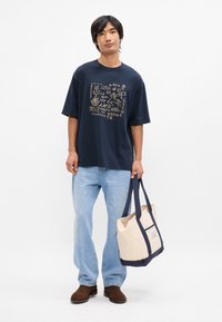 Ναυτικό βαμβακερό t-shirt με χρυσό γραφικό σχέδιο, χαλαρή εφαρμογή. Ανοιχτό μπλε τζιν παντελόνι, καφέ σουέτ υποδήματα και μπεζ τσάντα με ναυτικές λεπτομέρειες.