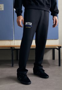 Crne trenirke s "INTER" logotipom u plavom i žutom tekstu s lijeve strane, uparene s crnim tenisicama. Tkanina izgleda mekano i teksturirano.