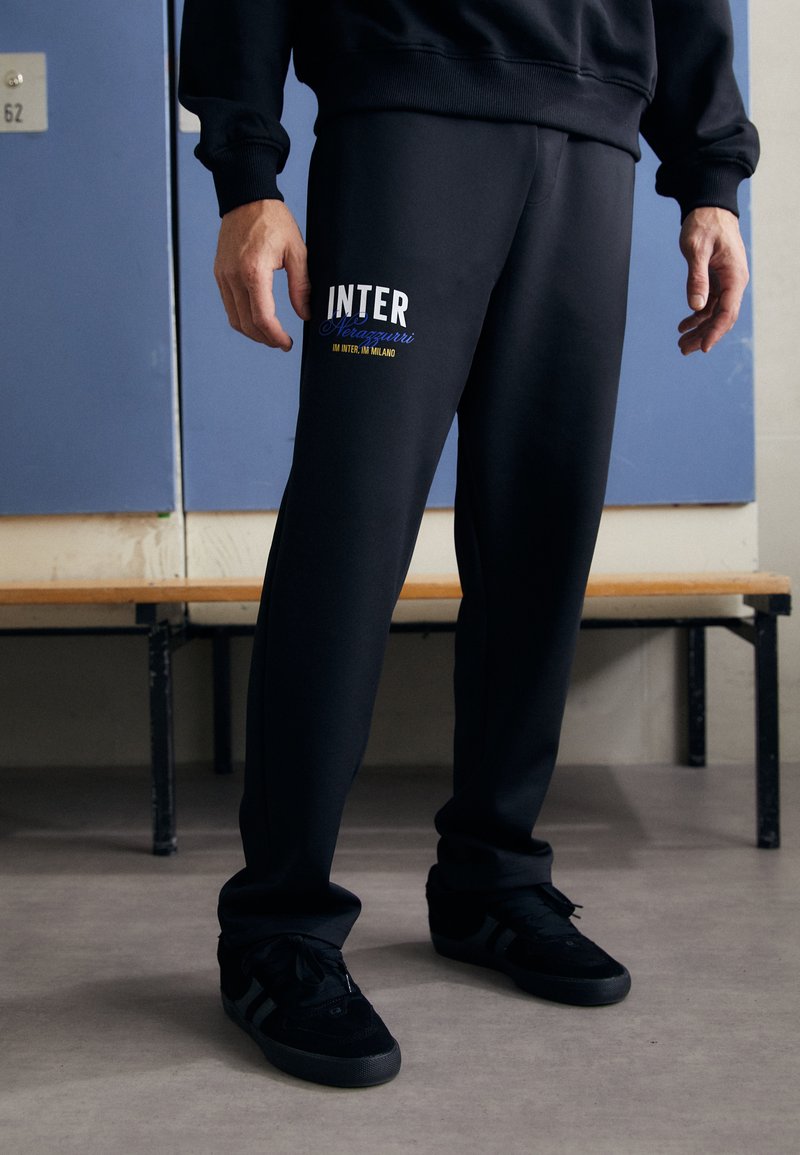 Crne trenirke s "INTER" logotipom u plavom i žutom tekstu s lijeve strane, uparene s crnim tenisicama. Tkanina izgleda mekano i teksturirano.