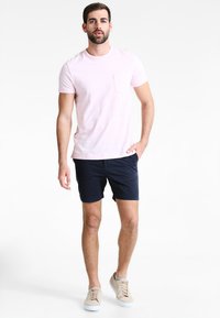 Lyserød kortærmet t-shirt med brystlomme, parret med marineblå shorts og beige sneakers. Glat stofstruktur, afslappet pasform.