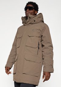 Brun, isoleret parka med høj krave, flere lommer foran og lynlås. Har hætte og justerbare manchetter.