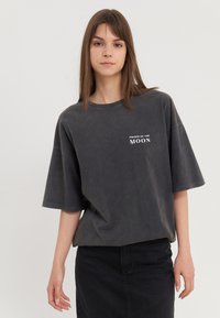 Dark grey oversized t-shirt, short sleeves, crew neckline, features white text "PHASES OF THE MOON" on the left chest area, slightly textured fabric.