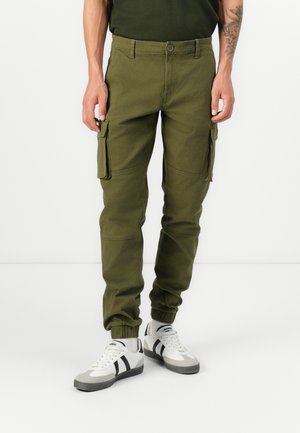 Man draagt olijfgroene cargobroek met zijzakken en witte sneakers met zwarte strepen, staande tegen een witte achtergrond.