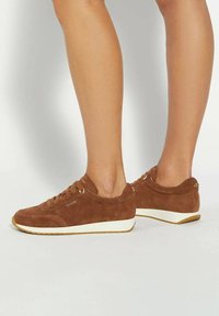 Benen in bruine suède veterschoenen met witte zolen, staande op een lichte achtergrond.