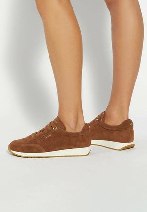 Benen in bruine suède veterschoenen met witte zolen, staande op een lichte achtergrond.