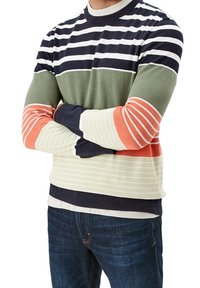 Mann mit verschränkten Armen, der einen gestreiften Pullover in Navy, Grün, Orange und Creme sowie eine dunkle Jeans trägt, von Hals bis Hüfte gezeigt.