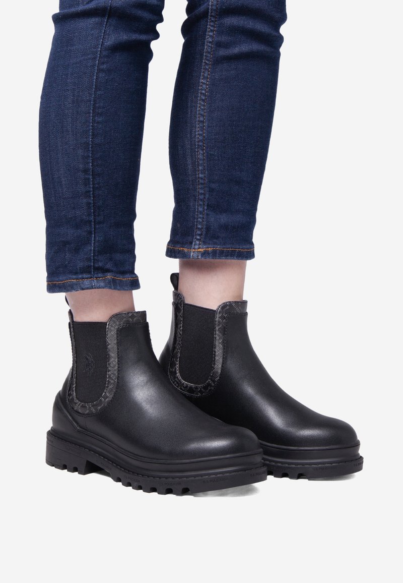 Černé kotníkové boty s elastickými bočními panely, texturovanými detaily a robustními gumovými podrážkami. V kombinaci s džíny, vytvářejícími uvolněný vzhled.