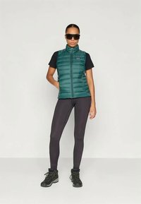 Γυναίκα που φοράει μαύρα γυαλιά ηλίου, πράσινο επενδυμένο γιλέκο, μαύρο κοντομάνικο πουκάμισο, μαύρα κολάν και μαύρες ορειβατικές μπότες, σε όρθια θέση σε λευκό φόντο.