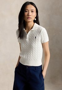 Young woman with long dark hair wears white cable-knit polo shirt and navy blue pants, standing with hands in pockets against plain background.