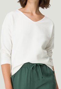 Mujer con una camiseta blanca de manga larga con textura metida dentro de pantalones verde oscuro con cordón, de pie contra un fondo liso.