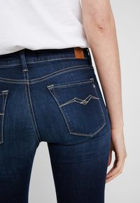 Jeans de denim azul oscuro con un corte ajustado, que cuentan con dos bolsillos traseros con costuras distintivas y un parche de cuero marrón con el logo en la cintura.