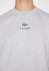 T-shirt grigia a girocollo realizzata in cotone, con un logo di coccodrillo ricamato in verde e la parola "LACOSTE" in verde sotto il logo.