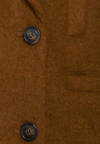 Brun uldfrakke vist tæt på, med en blød tekstur, to mørke knapper og en sidelomme; overordnet ensfarvet med minimal detaljering.
