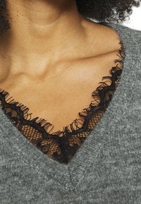 Szary sweter dzianinowy z dekoltem w kształcie litery V, podkreślony czarną koronką wzdłuż dekoltu, eksponujący delikatną teksturę i kontrast.