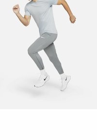 Mann in grauer Sportbekleidung und weißen Nike-Sneakern, der im Lauf gegen einen schlichten weißen Hintergrund läuft.